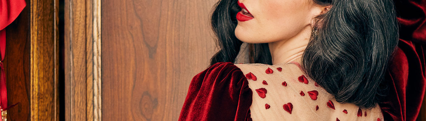 Close up of model in glamorous curls worn with red lipstick, model wears red velvet bed jacket with red heart embroidery over the shoulders for Valentine's Day style.