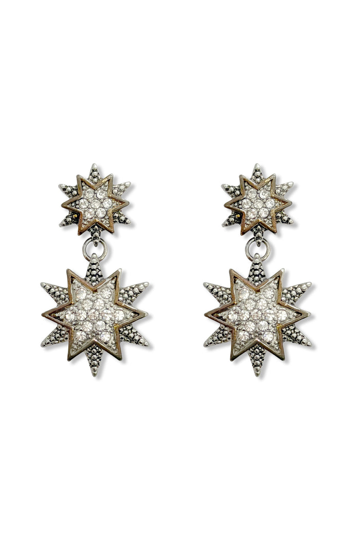 A pair of star-shaped drop earrings with pavé-set clear stones with gold-toned and silver-toned metal borders. These six-pointed stars are Victorian empress style and glamorous for showgirl fashion, shown on a plain white background